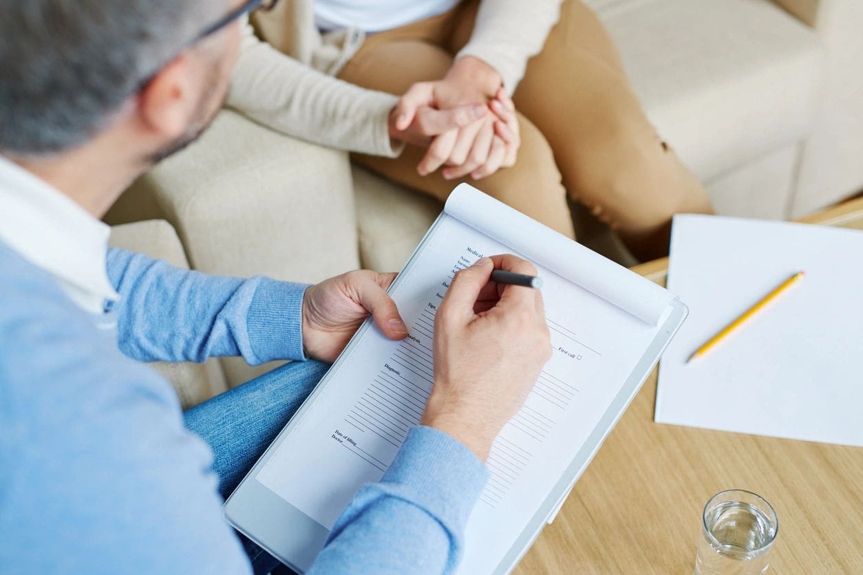 A man writing on paper while sitting next to another person.